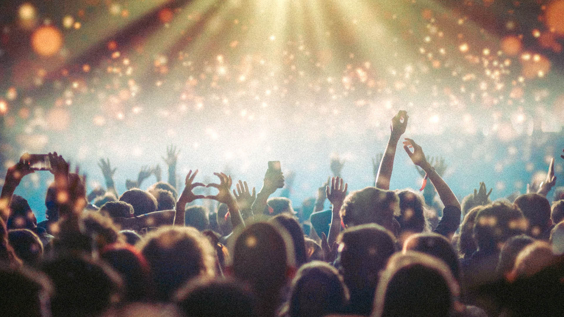 Menschenmenge auf einem Konzert, viele jubelnde Personen mit erhobenen Armen vor einer hell beleuchteten Bühne, darüber schwebende, unscharfe Lichtpunkte, die eine festliche, energetische Stimmung erzeugen.